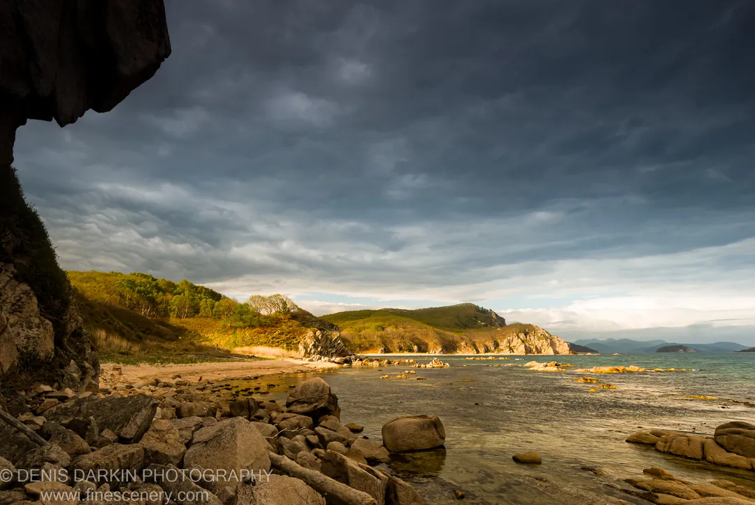 A dark rocky overhang at Petrova Island with rolling green hills lit by low-angle sunlight visible beyond the calm water.