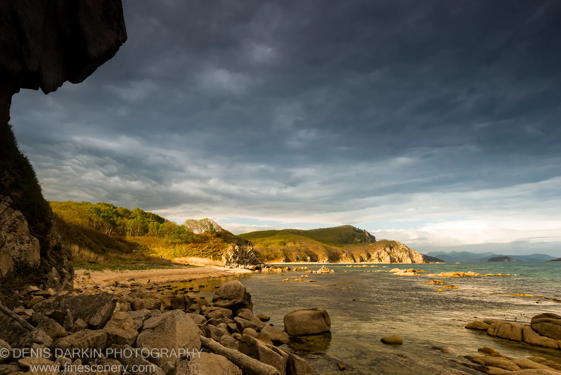 A dark rocky overhang at Petrova Island with rolling green hills lit by low-angle sunlight visible beyond the calm water.