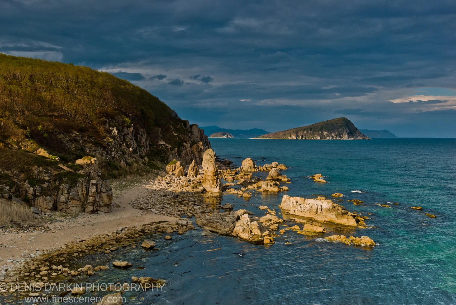 Warm light on limestone boulders along a pebble beach at Petrova Island, with a large island and dark clouds on the horizon.
