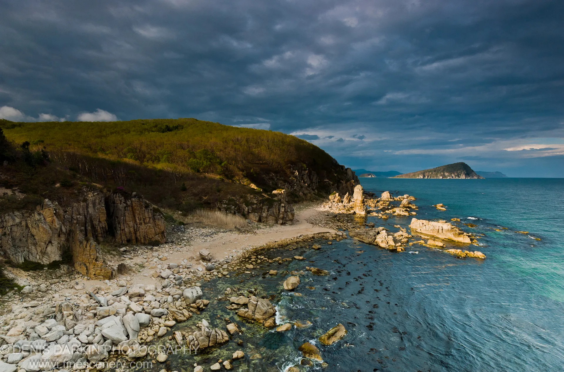 A sheltered pebble cove at Petrova Island lined with columnar basalt, with turquoise water and a storm-clouded sky overhead.