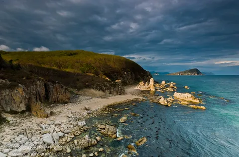 A sheltered pebble cove at Petrova Island lined with columnar basalt, with turquoise water and a storm-clouded sky overhead.