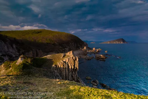 Sunlit sea cliffs with vertical rock striations at Petrova Island, with green slopes meeting deep blue water and an offshore island on the horizon.