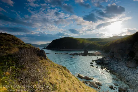 Rolling green hills enclosing a sheltered bay on Petrova Island, Lazovsky Reserve, with deep blue water and scattered cumulus clouds above.