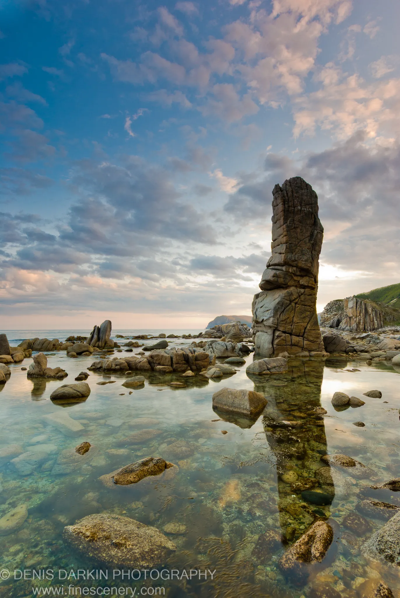 A tall stratified rock pillar reflected in the glassy shallow water of a tidal cove at Petrova Island at dusk.
