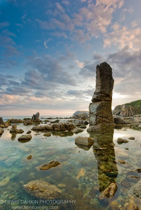 A tall stratified rock pillar reflected in the glassy shallow water of a tidal cove at Petrova Island at dusk.