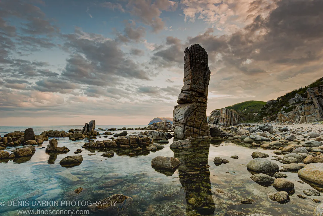 A sea stack towers over a glass-like tidal pool at Petrova Island, with soft dusk light catching the clear water and distant hills.