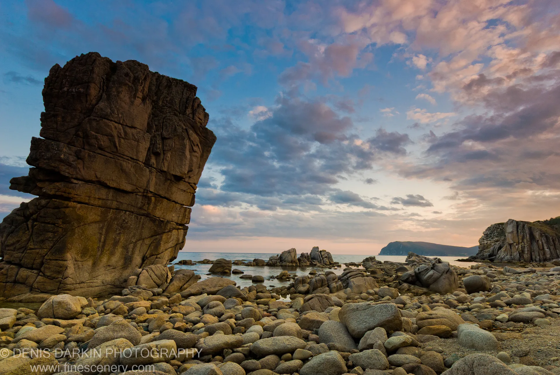 A massive layered rock stack stands beside calm water at Petrova Island, with a twilight sky of pink and blue above the pebble shore.