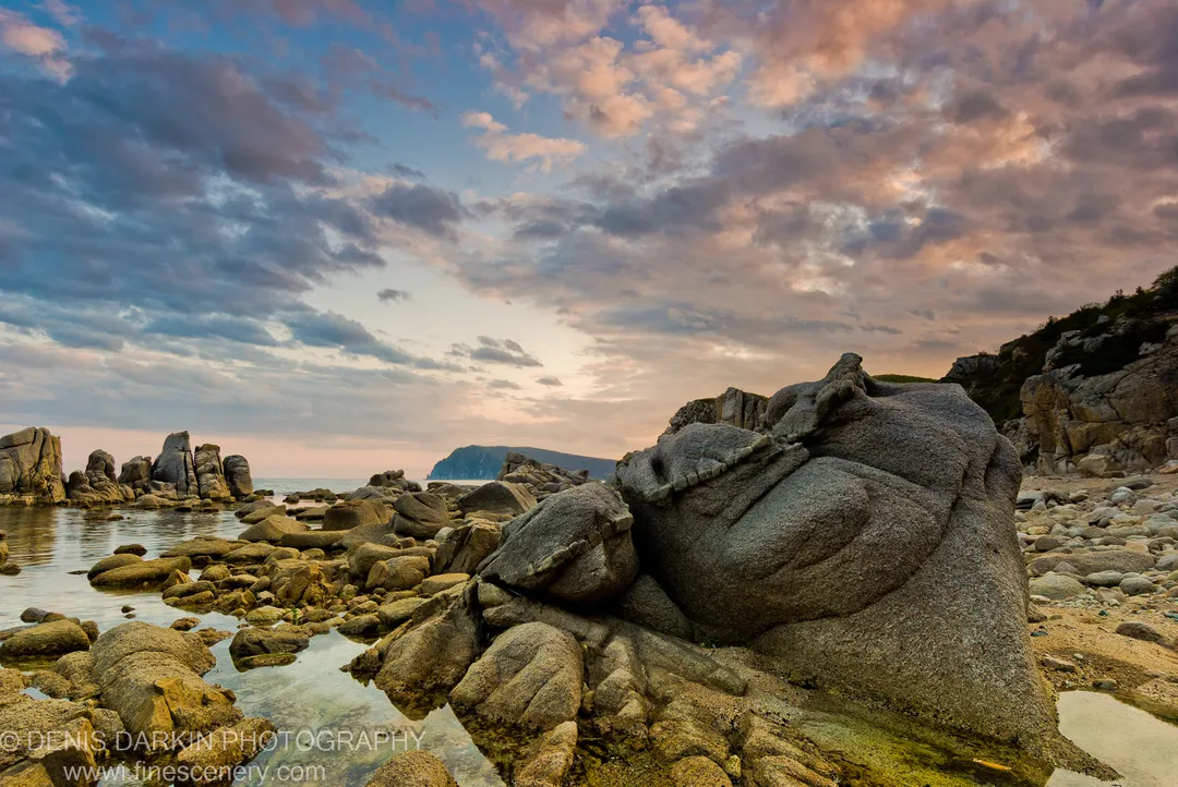 Smooth granite boulders on the shore of Petrova Island at dusk, shallow pools reflecting a gradient of pink and deep blue in the sky.