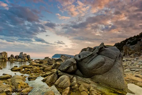 Smooth granite boulders on the shore of Petrova Island at dusk, shallow pools reflecting a gradient of pink and deep blue in the sky.