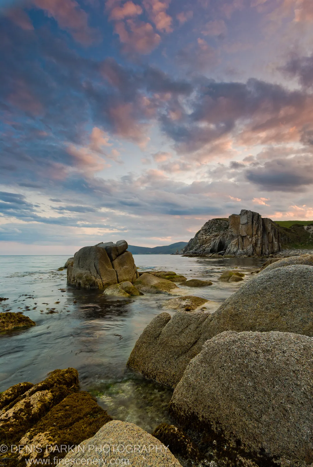 Moss-covered granite boulders at Petrova Island with pink and purple clouds at dusk, the calm sea stretching to a cliff face.