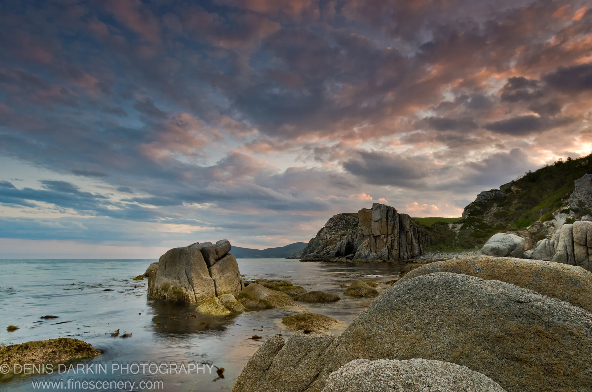 Moss-covered granite boulders rest in still water at the Petrova Island headland, beneath a sky moving from deep blue to pink at evening.