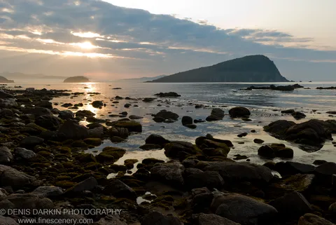 Moss-covered boulders line the foreground of a calm bay on Petrova Island, with sunlight breaking through clouds toward a forested headland.
