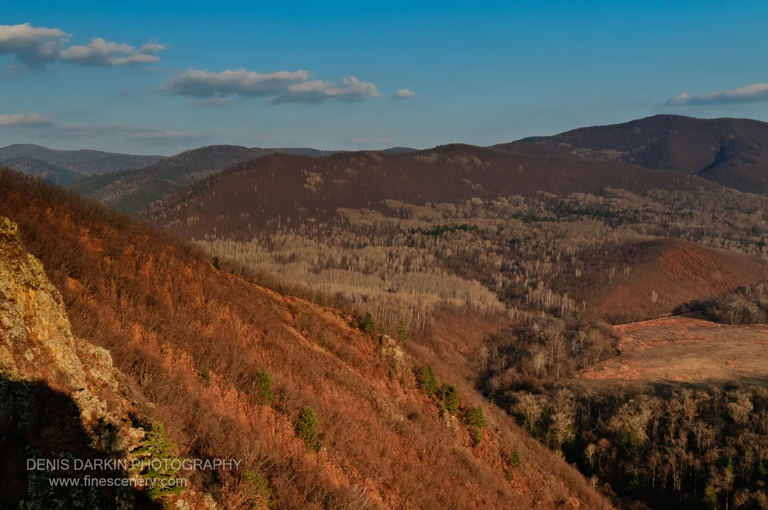A moss-covered rock face overlooks the Izvilinka River valley, where bare deciduous trees mix with evergreen forest above distant blue ridges.