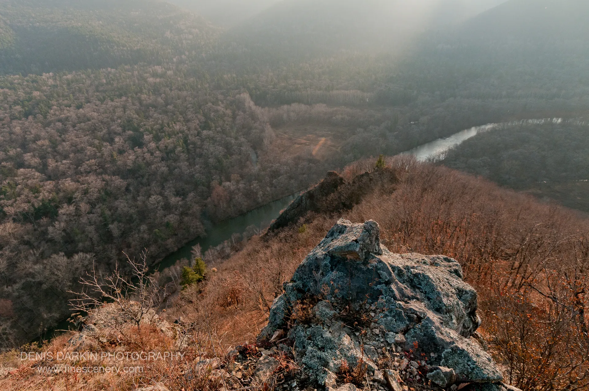 A lichen-covered rock on a ridge overlooks the Izvilinka River winding through a haze-filled valley of dormant forest.