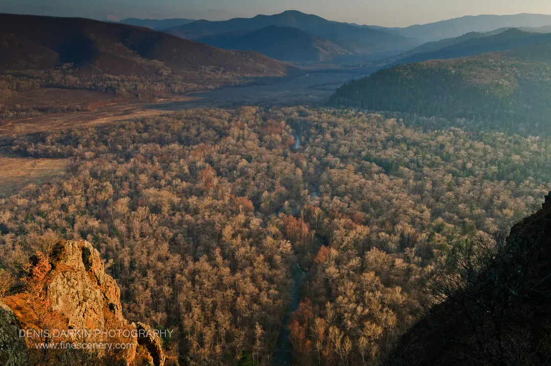 Warm-lit rock face in the foreground contrasts with the cool blue haze of the Izvilinka River valley and distant mountain ridges.