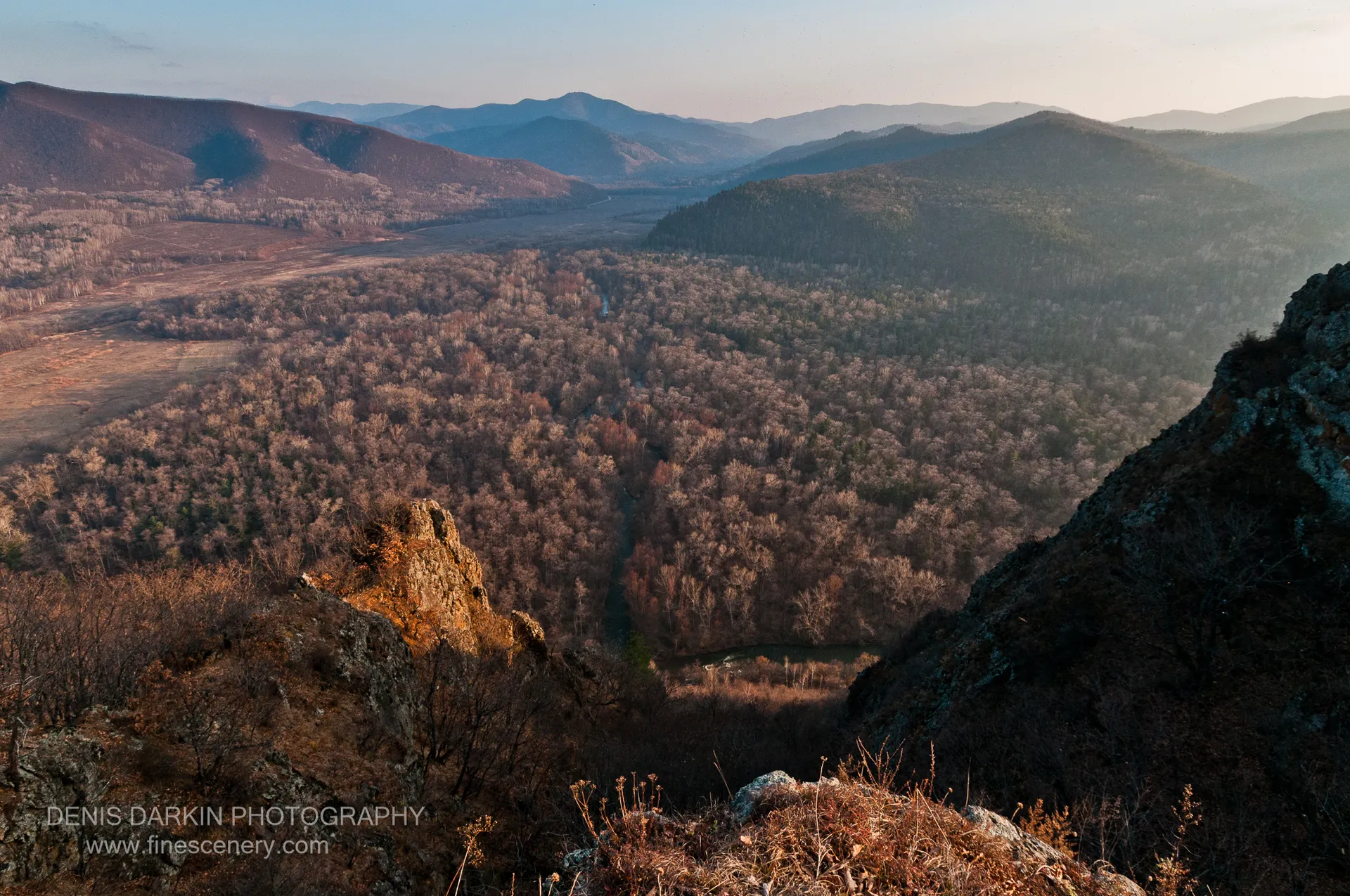 Golden light on rugged foreground rocks contrasts with the cool shadows of distant mountain layers over a winding river valley in Chuguyevsky District.