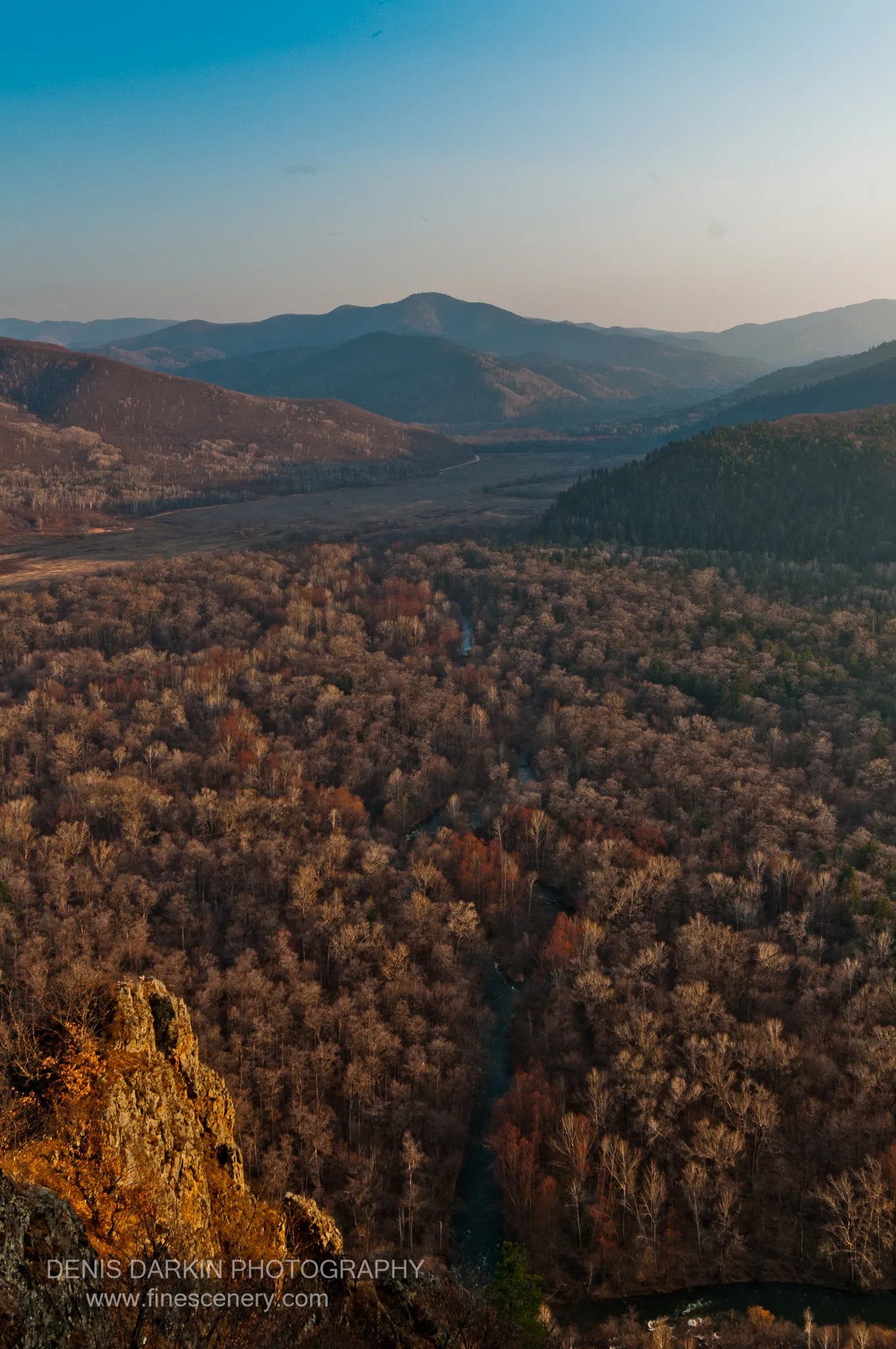 A rocky cliff edge in late afternoon light overlooks a deep valley where the river winds through bare and russet-toned trees.