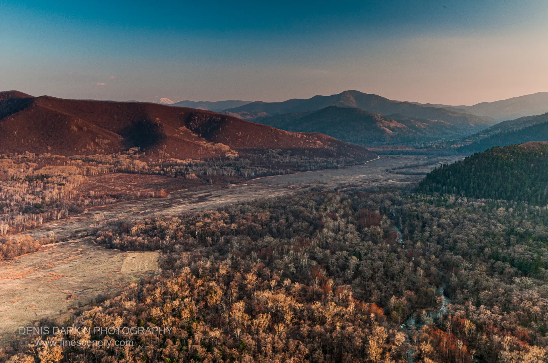 Warm golden-hour light on a mixed forest canopy above the Izvilinka gravel riverbed, with layered mountain ranges receding into blue haze.