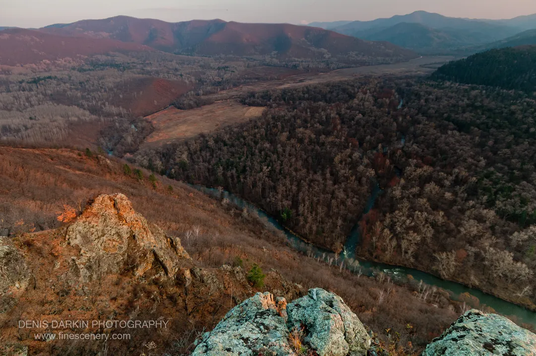 Warm afternoon sunlight on lichen-covered rocks above the Izvilinka River, with cool blue tones in the water and distant ridgelines.