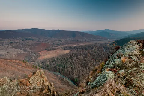 Lichen-covered boulders and bare trees beside a winding river, with layered mountains receding into a sunset sky, Izvilinka River.