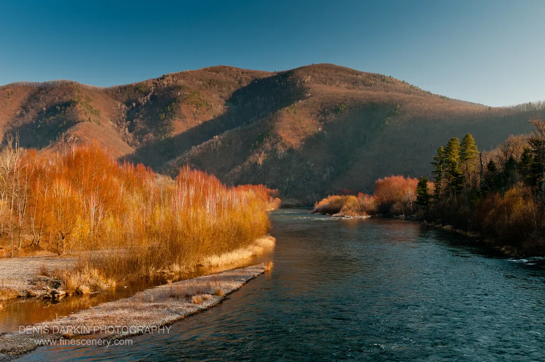 Wide river past a frosted gravel shore and mixed autumn forest of birch and conifer, mountain ridgeline behind, Ussuri River.