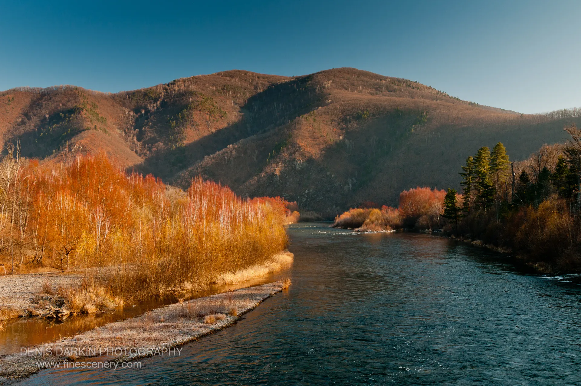 Wide river past a frosted gravel shore and mixed autumn forest of birch and conifer, mountain ridgeline behind, Ussuri River.
