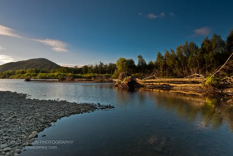 Grey stones on the left bank of the Bikin River lead toward downed timber and dense green forest along the right shore.
