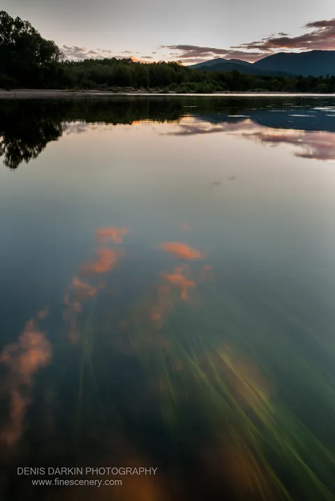 The Bikin River at dusk mirrors scattered orange clouds and mountain silhouettes in its glassy surface, submerged vegetation visible in the clear shallows below.