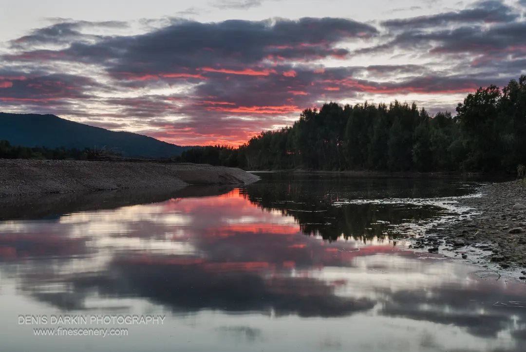 Rose and violet cloud streaks reflected in the Bikin River at dusk, rough pebbles in the foreground and dark forest at right.