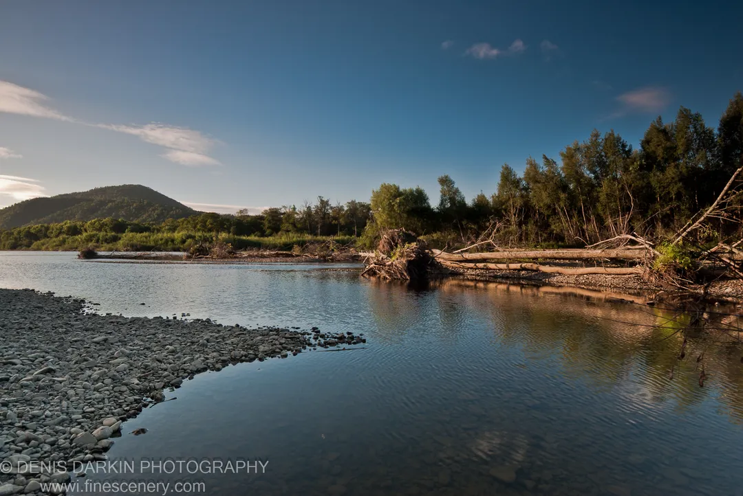 Pebbled Bikin River shore in morning, submerged stones visible and a forested hill beyond the clear water under pale blue sky.