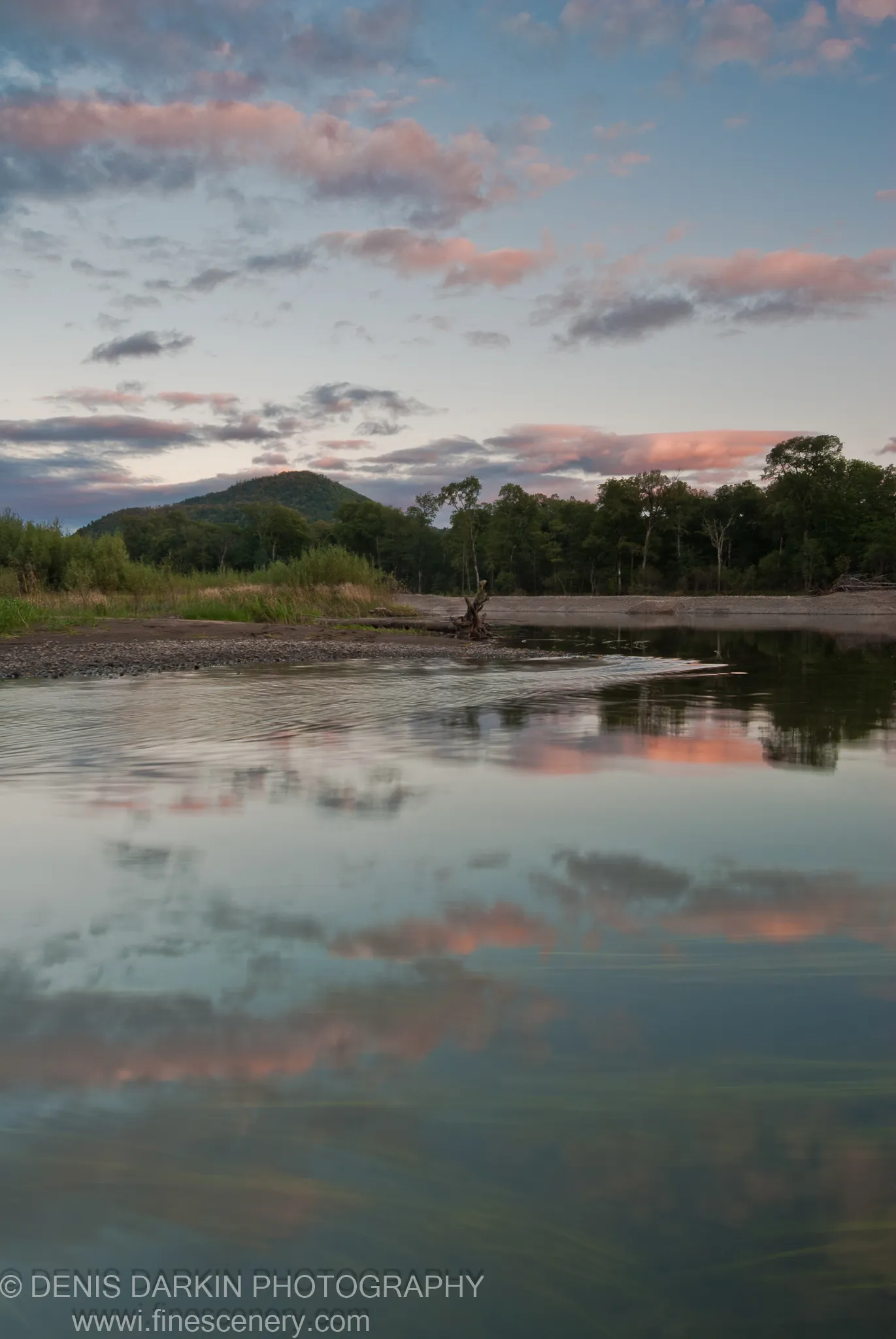 The Bikin River at dusk with a forested hill reflected in calm water, pink-tinted clouds above and a gravel bank in the left foreground.