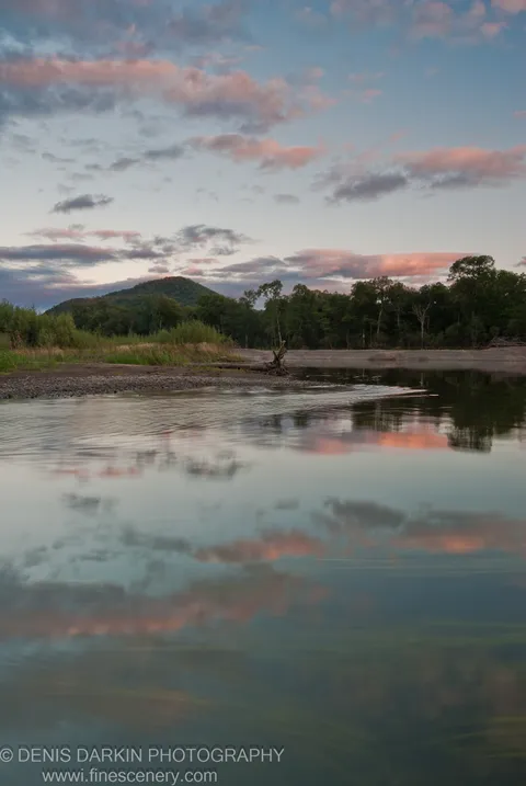 The Bikin River at dusk with a forested hill reflected in calm water, pink-tinted clouds above and a gravel bank in the left foreground.