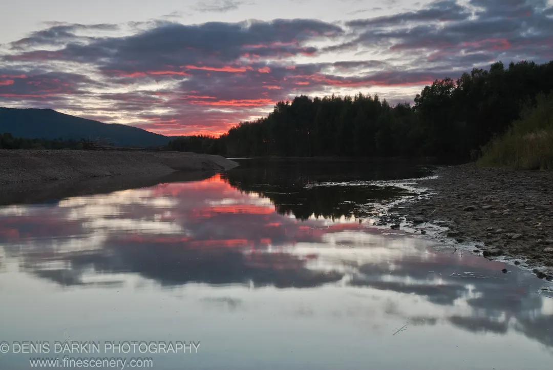 Calm water on the Bikin River at dusk reflects pink and purple clouds, a silhouetted forest on the horizon and a pebbly bank at right.