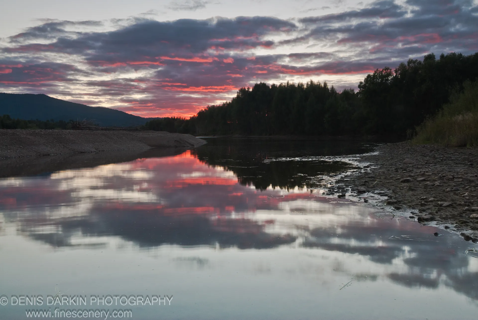 Calm water on the Bikin River at dusk reflects pink and purple clouds, a silhouetted forest on the horizon and a pebbly bank at right.
