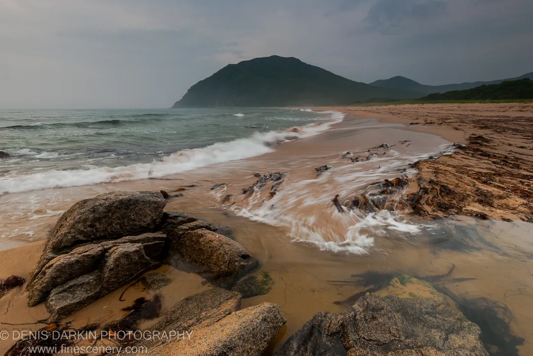 Dark, textured boulders and scattered driftwood anchor the foreground of a wide-angle morning seascape at Tachingouza Bay, with a forested mountain under overcast skies.