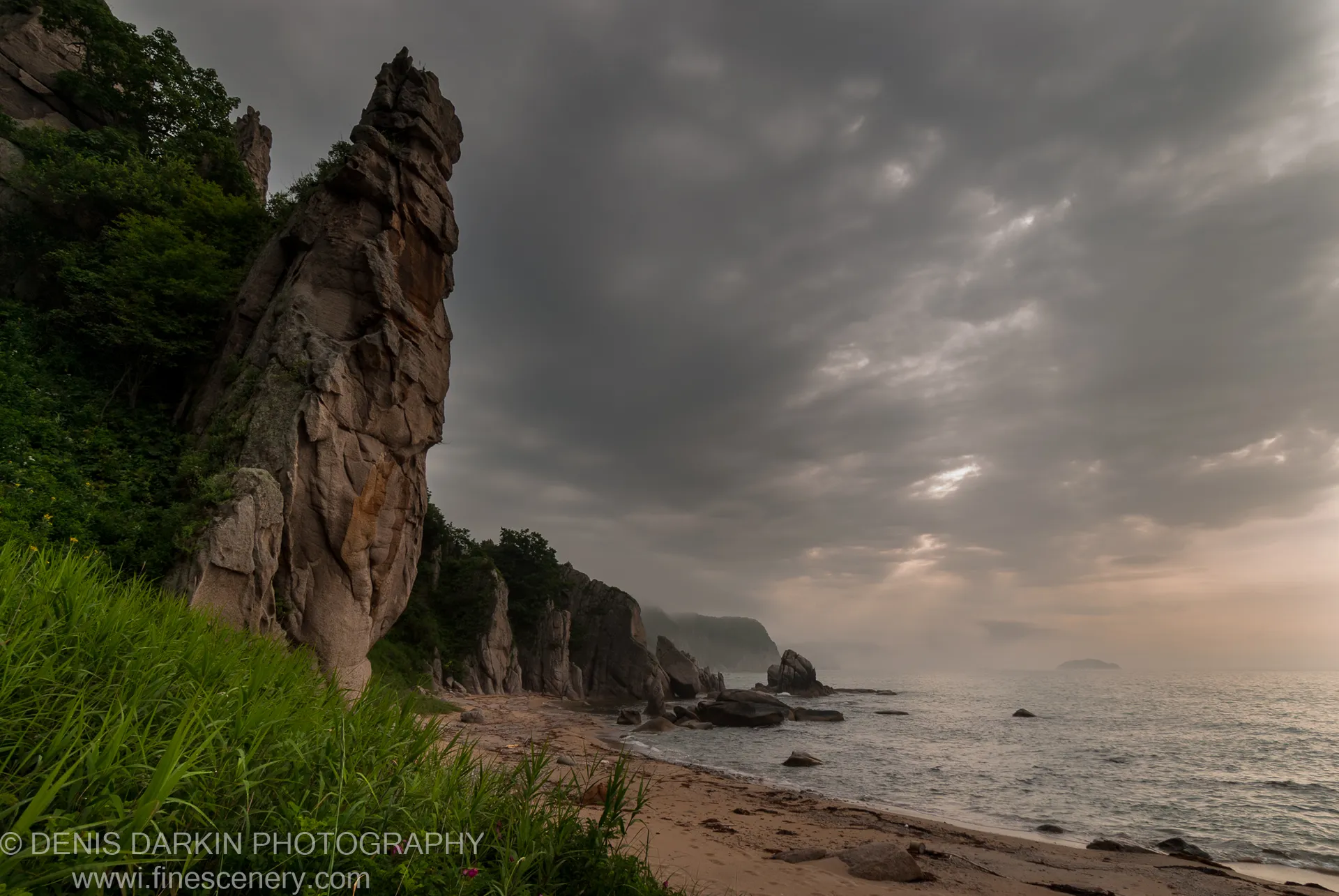 A weathered rock pinnacle rises from dense coastal vegetation beside a curved sandy beach under heavy grey overcast sky, Tachingouza Bay, July 2010.