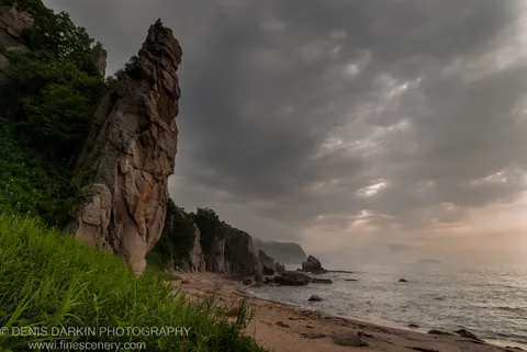 A weathered rock pinnacle rises from dense coastal vegetation beside a curved sandy beach under heavy grey overcast sky, Tachingouza Bay, July 2010.