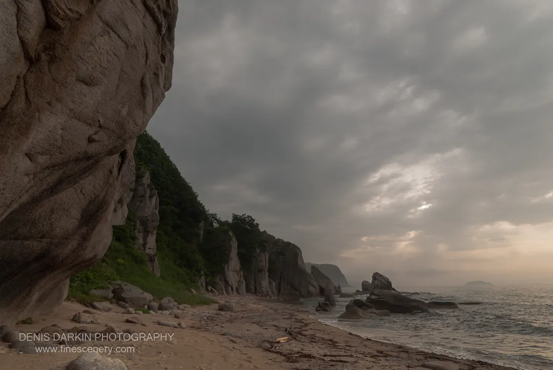 A massive rock formation stands at the left of the frame, with a sandy beach curving past sea stacks along the Lazovsky coast under overcast skies.