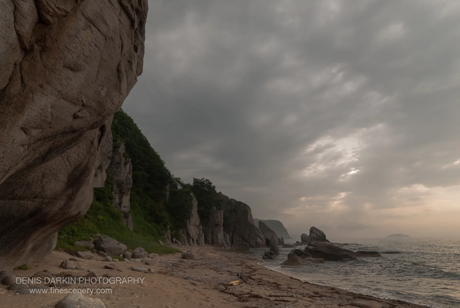 A massive rock formation stands at the left of the frame, with a sandy beach curving past sea stacks along the Lazovsky coast under overcast skies.