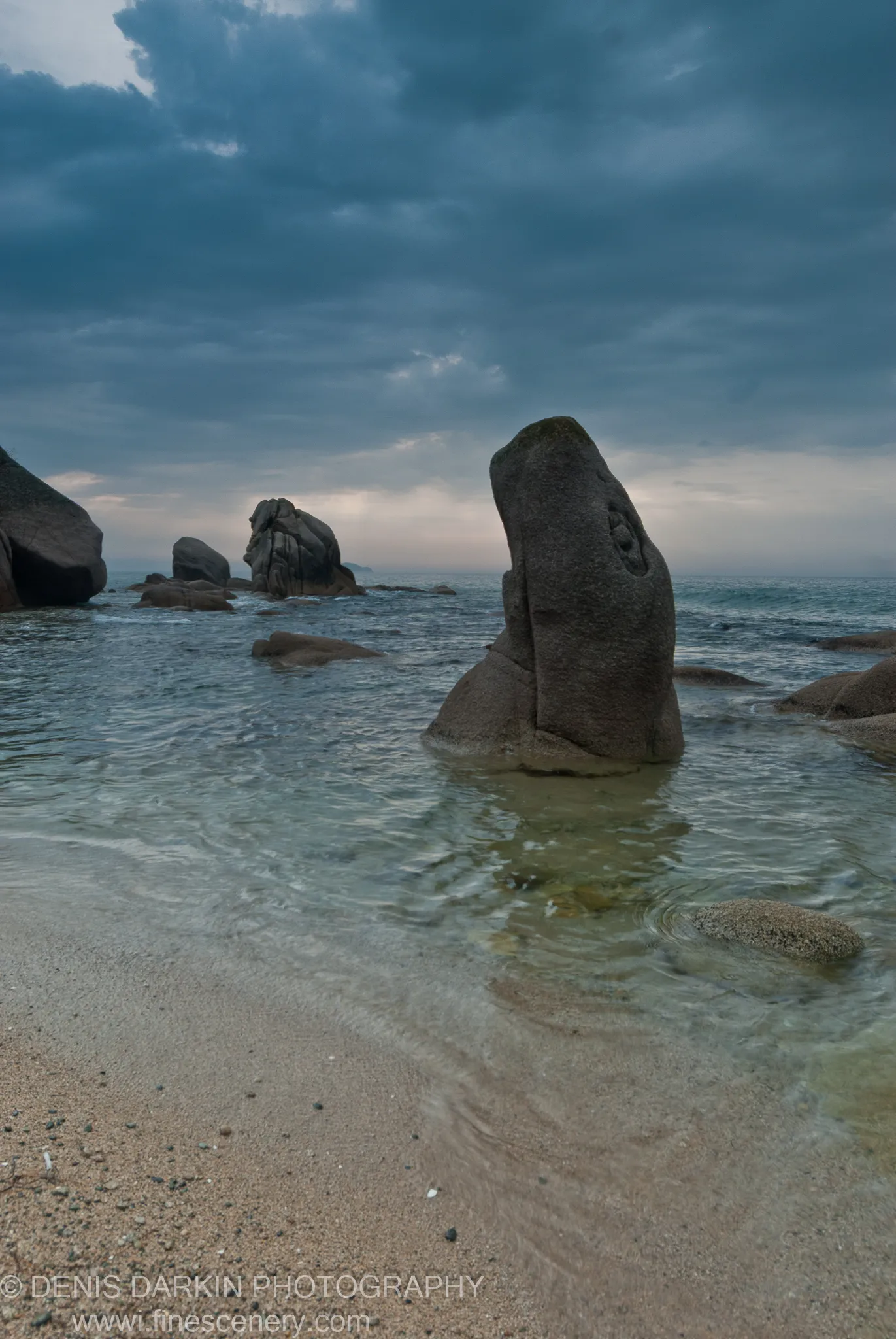 Dark clouds hang over weathered granite rocks rising from clear, shallow water at Tachingouza Bay, with a sandy beach and textured stone in the foreground.