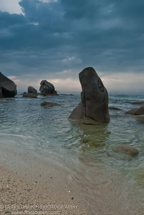 Dark clouds hang over weathered granite rocks rising from clear, shallow water at Tachingouza Bay, with a sandy beach and textured stone in the foreground.