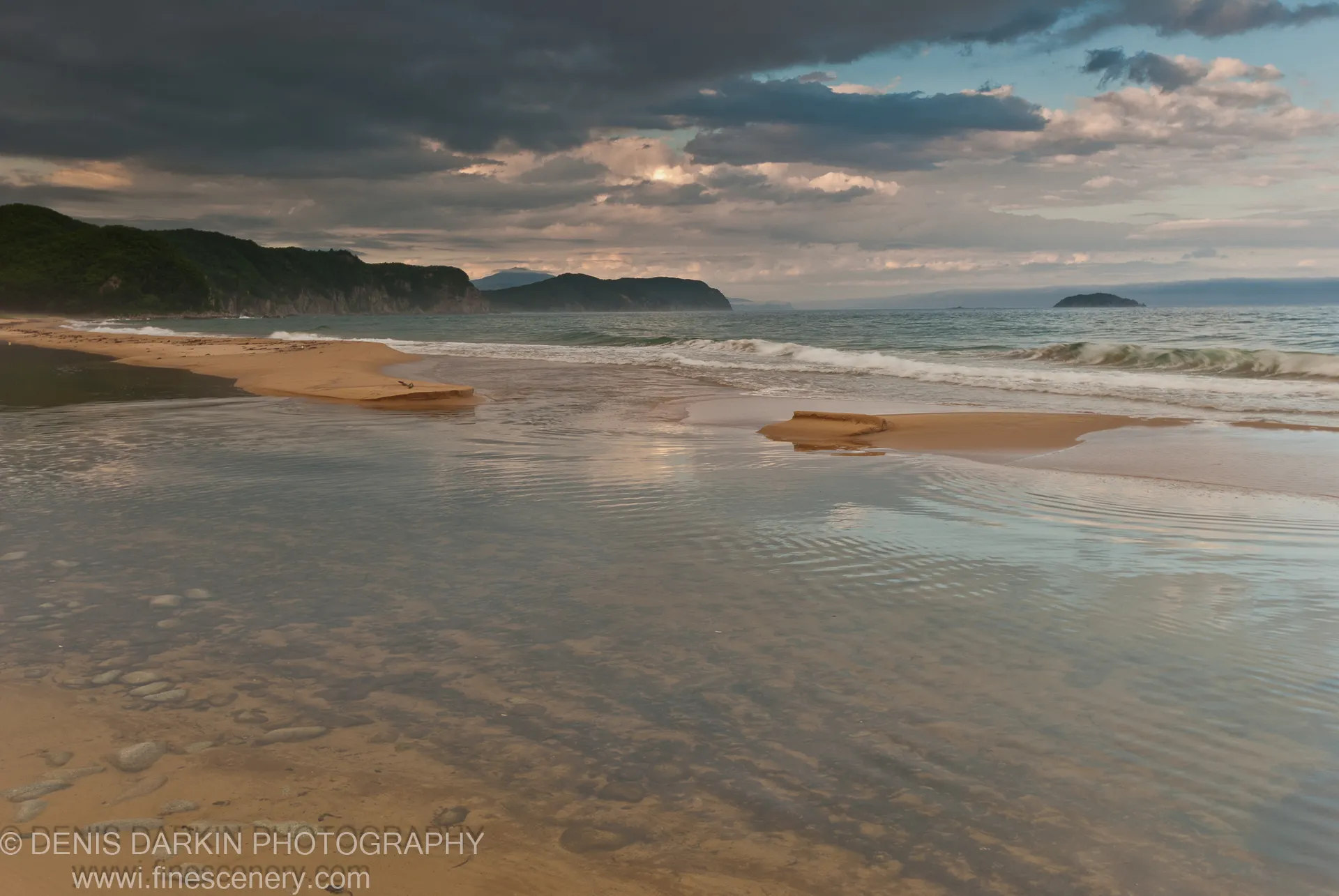 Two sandy spits extend into the ocean at dusk, with submerged stones in the foreground and storm clouds lit by golden sunlight above dark headlands.