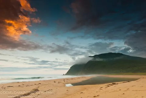 Fog-veiled coastal hills rise above a sandy beach and creek mouth at evening, with warm clouds on the left and cool blue water below.