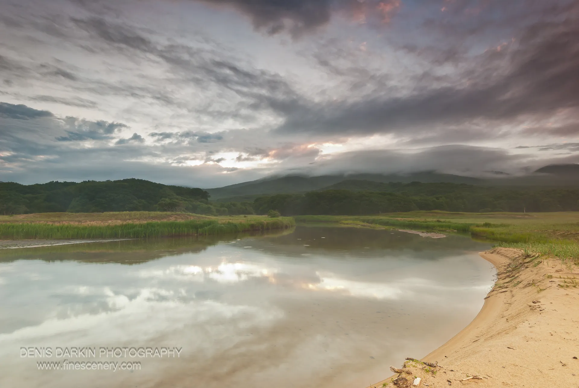A wide river curve at Tachingouza Bay mirrors a stormy sky at evening, with mist-covered forested hills creating atmospheric depth in the background.