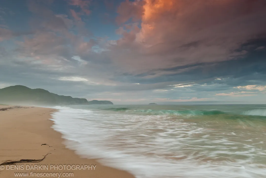 Long-exposure waves roll onto a sandy beach at sunset, with a large cloud formation catching warm orange light above the bay at Tachingouza.