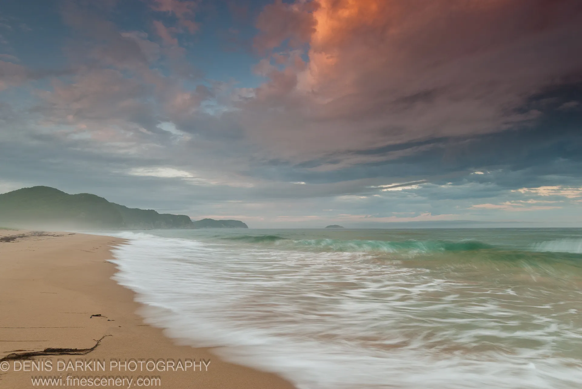 Long-exposure waves roll onto a sandy beach at sunset, with a large cloud formation catching warm orange light above the bay at Tachingouza.