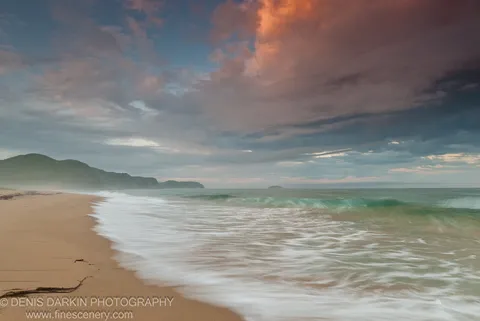 Long-exposure waves roll onto a sandy beach at sunset, with a large cloud formation catching warm orange light above the bay at Tachingouza.