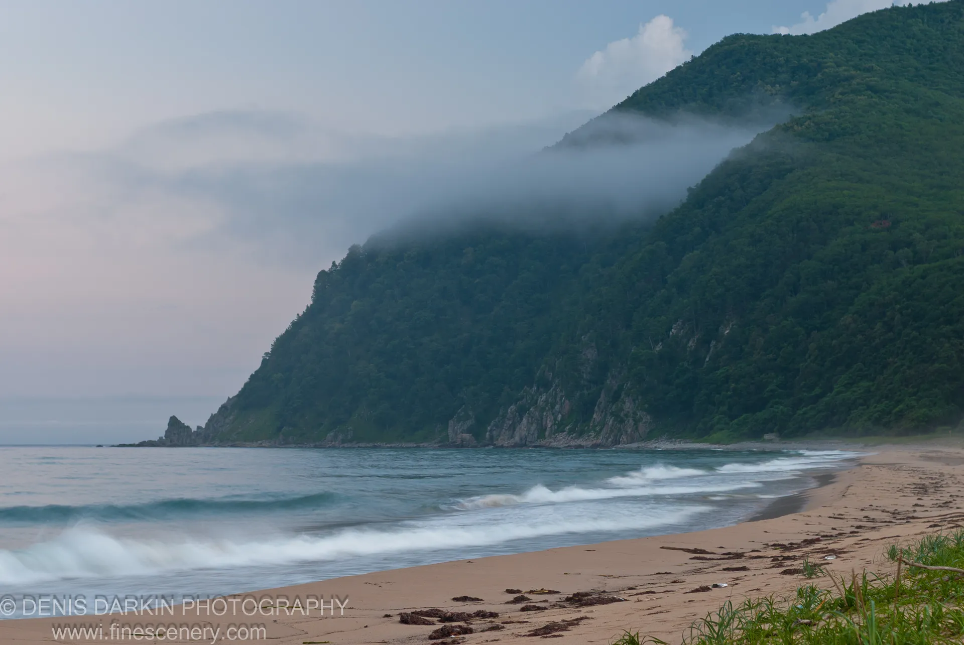 A fog-shrouded forested mountain slopes to a sandy beach at Tachingouza Bay, with gentle breaking waves and scattered dark seaweed under an overcast sky.