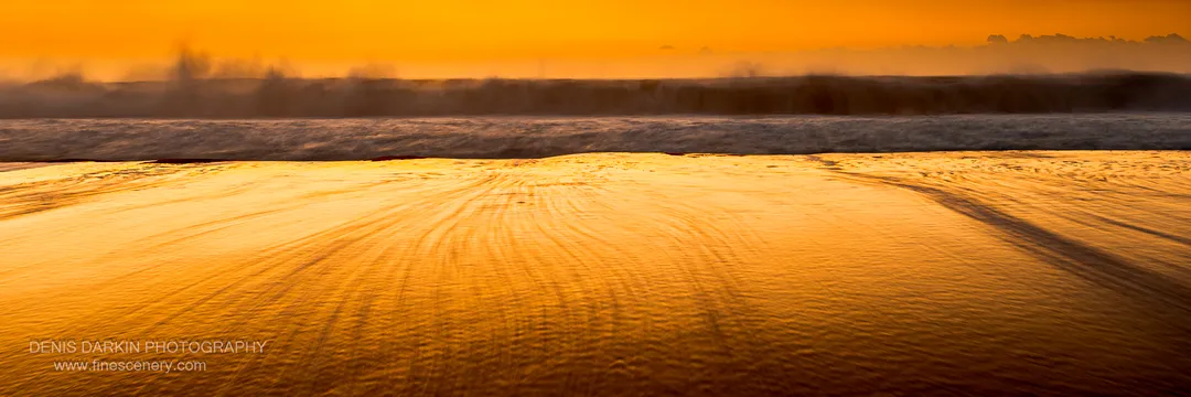 Rippled wet sand reflecting intense orange light at dawn on Spoon Bay beach, with a misty horizon beyond breaking waves.