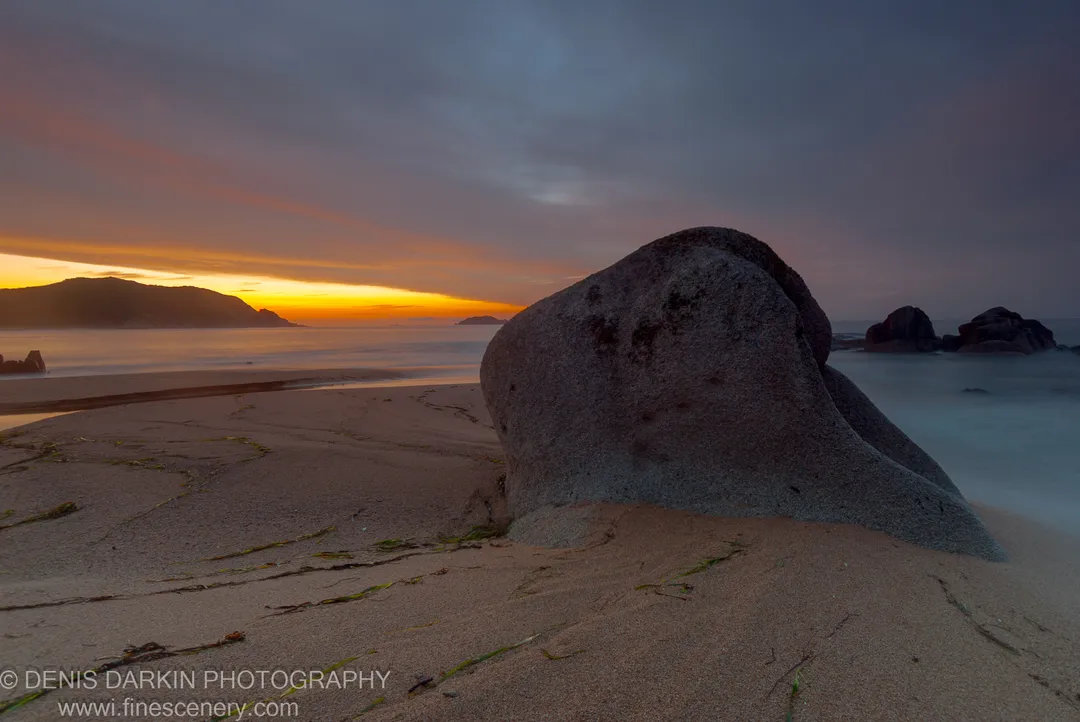 A large boulder on the sandy beach at Tachingouza Bay, with long-exposure surf as mist and a warm orange horizon at dawn.